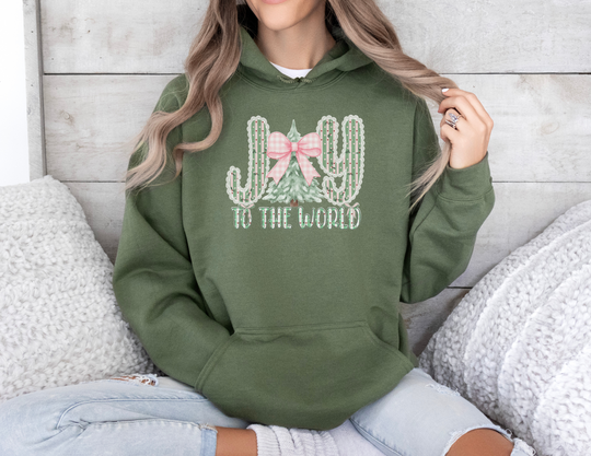 Person in forest-green JOY to the World hoodie with cactus and tree graphic, sitting on a light couch with chunky knit pillows, wearing ripped jeans.