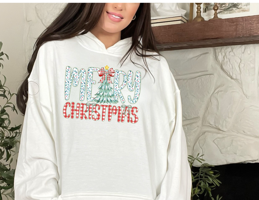 Woman wearing Tree Christmas Hoodie with MERRY CHRISTMAS graphic in holly-leaf and gingham fonts; cozy cotton-poly blend, featuring kangaroo pocket and matching drawstring.