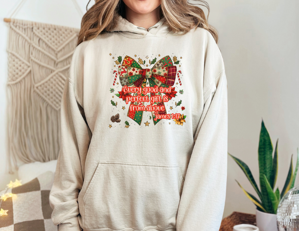 Person wearing Gift from Above Hoodie featuring a patchwork Christmas bow with holiday motifs and James 1:17 verse, against a cozy home décor background.