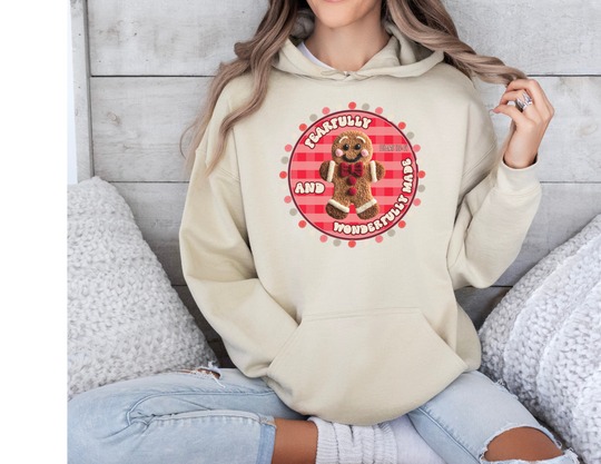 Person in light beige Wonderfully Made Hoodie featuring a gingham gingerbread design and biblical quote, paired with ripped jeans and white socks, seated on a couch.