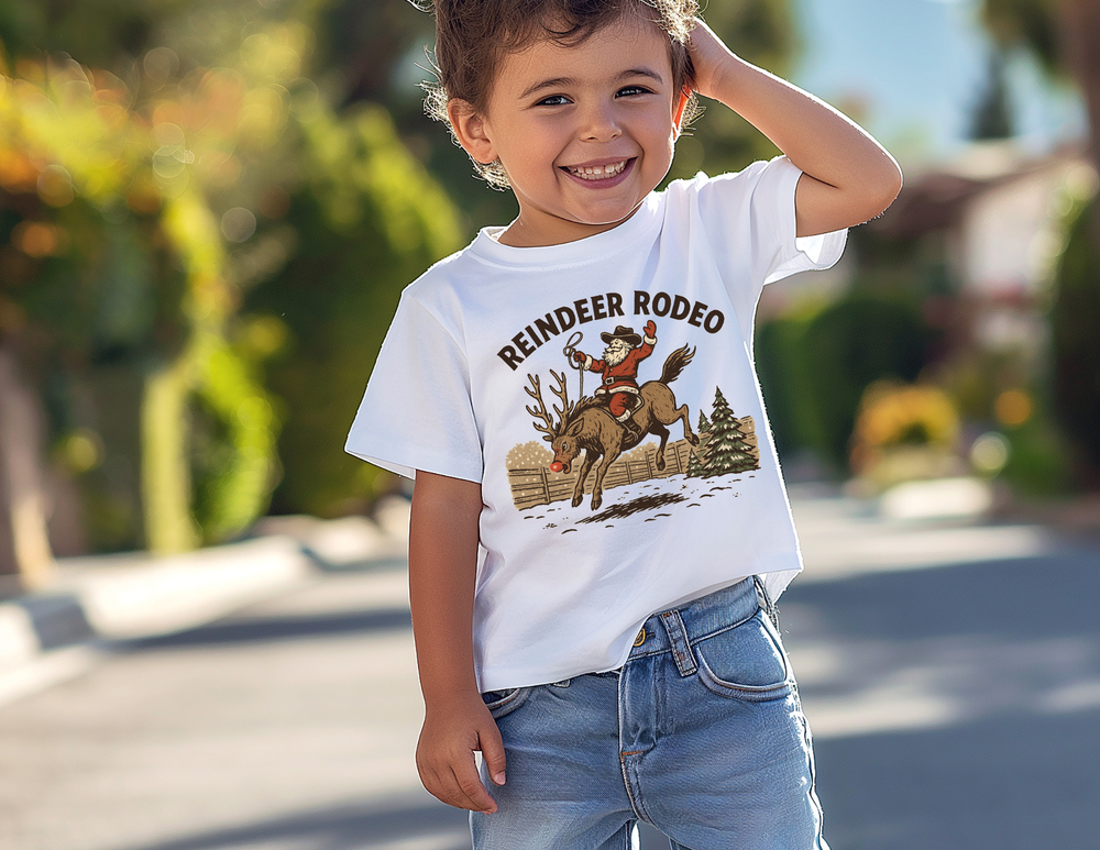 Toddler wearing Reindeer Rodeo Kids Tee with Santa on a reindeer, standing happily on a sunny street. Perfect for active play and comfort.