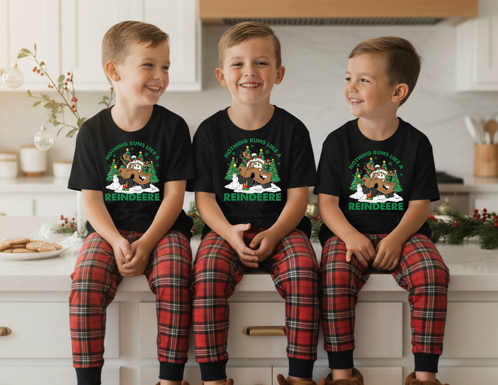 Three boys in matching Reindeer Kids Tees and plaid pajama pants sit on a kitchen island, showcasing festive holiday attire from Faith Family Designs.