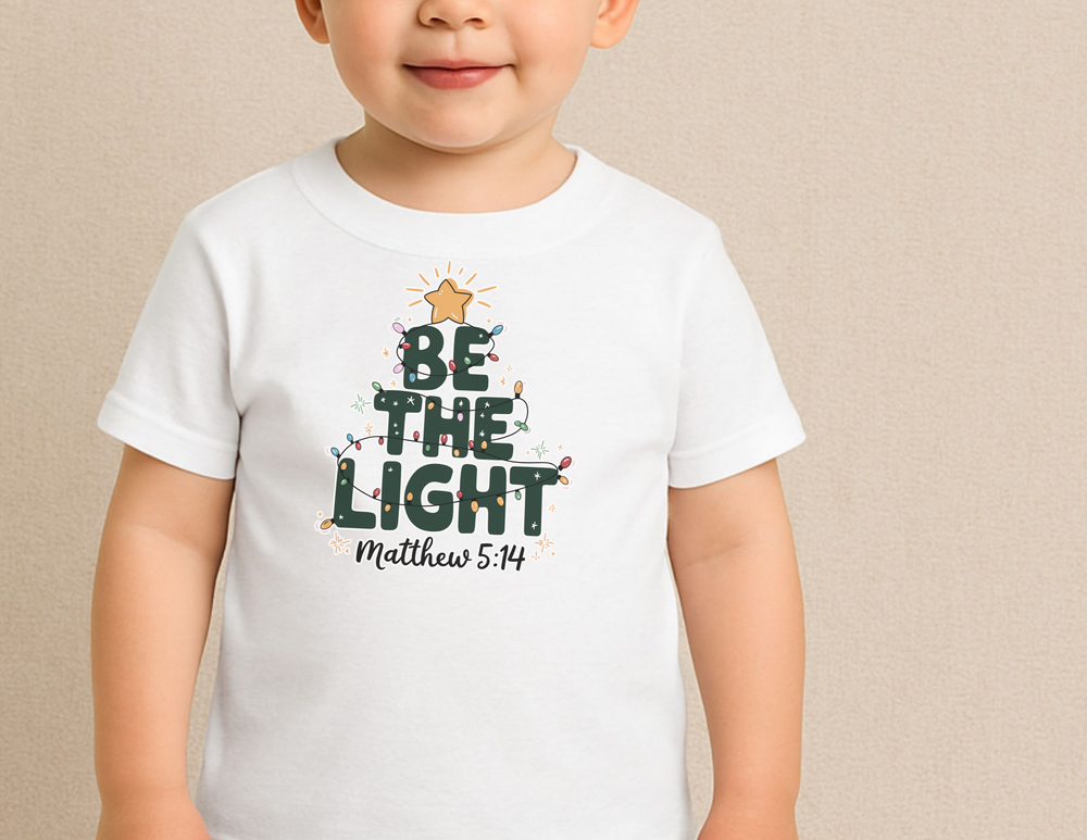 Child wearing Be The Light Toddler Tee with Christmas-tree graphic and Bible reference Matthew 5:14, designed for softness and durability.