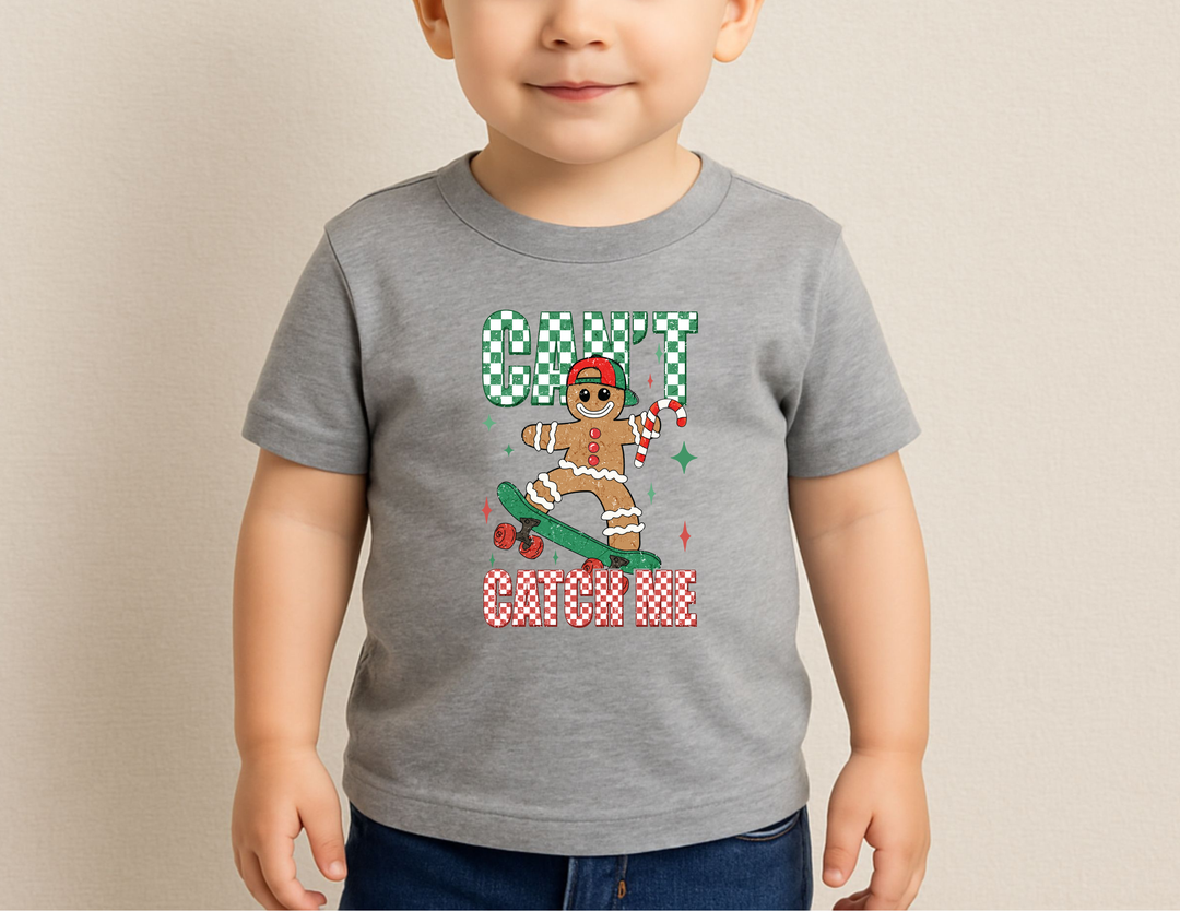 Can't Catch Me Toddler Tee featuring a cartoon gingerbread man on a skateboard, candy cane in hand, with playful red-green text. Perfectly soft for toddlers.
