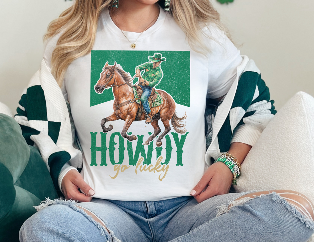 Howdy Go Lucky Tee featuring a cowboy on horseback, worn by a woman. This unisex jersey tee offers a soft cotton feel and durable fit.