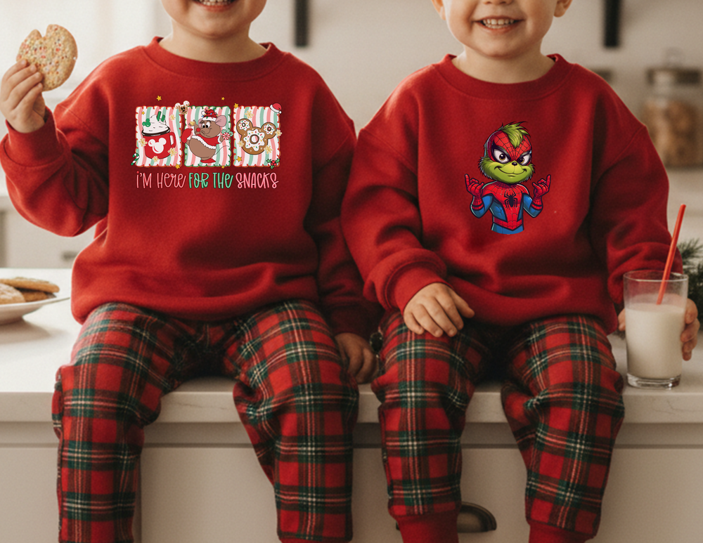 Two children wearing matching Here for the Snacks and Spider-Man-style sweatshirts, seated on a kitchen counter, enjoying a holiday moment with cookies and milk.
