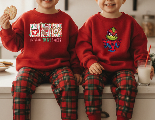 Two children wearing matching Here for the Snacks and Spider-Man-style sweatshirts, seated on a kitchen counter, enjoying a holiday moment with cookies and milk.
