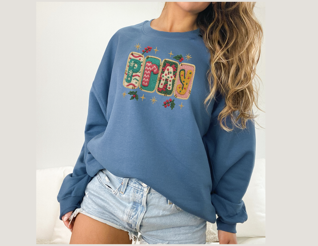 Person wearing a Pray Crew sweatshirt with festive applique, paired with light-wash denim shorts. The sweatshirt features colorful holiday-themed patches spelling PRAY.