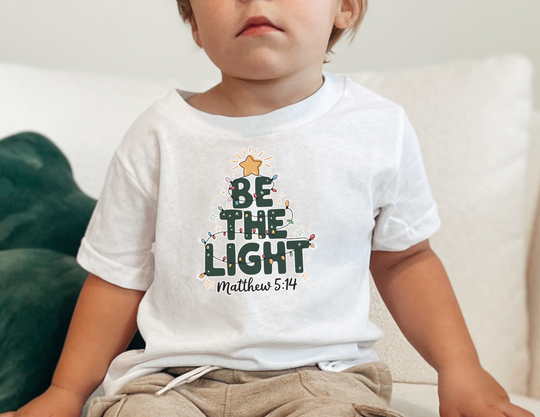 Child on couch wearing Be The Light baby tee with Christmas-tree graphic and Matthew 5:14 reference, showcasing Faith Family Designs' cozy, stylish, faith-inspired apparel.
