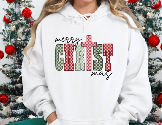 Person wearing a Merry CHRISTmas hoodie with a red and green checkerboard pattern, featuring a cross design. Christmas tree with red baubles in the background.
