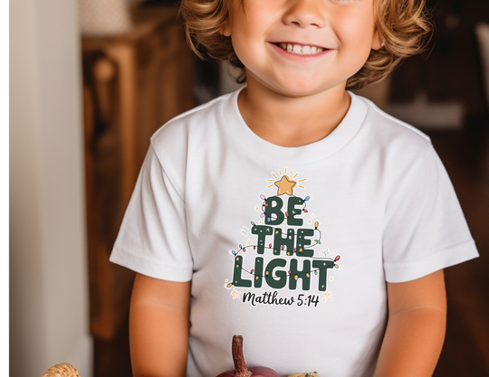 Child wearing Be The Light Kids Tee with Christmas-tree shaped text and Matthew 5:14 reference, highlighting the shirt's durable cotton fabric and timeless style.
