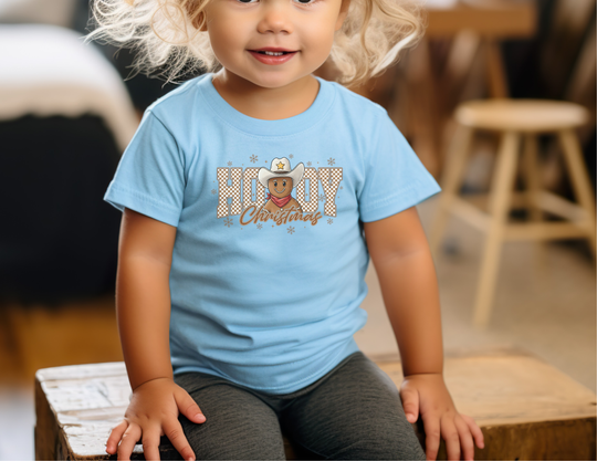 Young child wearing the Howdy Christmas Toddler Tee with a cartoon gingerbread cowboy design, seated on a wooden bench, showcasing the tee's softness and playful graphic.