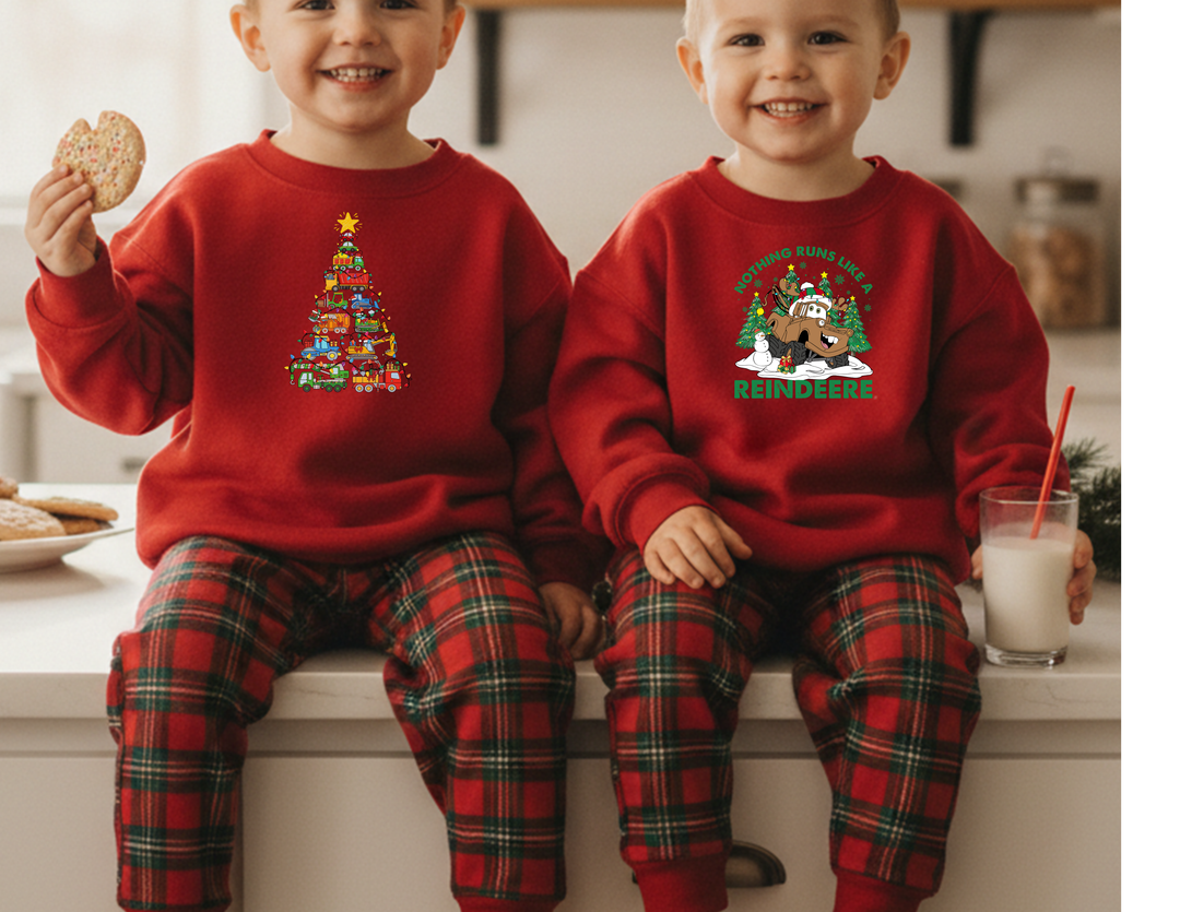 Two children sit on a kitchen counter in matching red Christmas Truck Toddler Crews, featuring festive tree and reindeer graphics, enjoying cookies and milk.