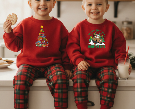 Two children sit on a kitchen counter in matching red Christmas Truck Toddler Crews, featuring festive tree and reindeer graphics, enjoying cookies and milk.