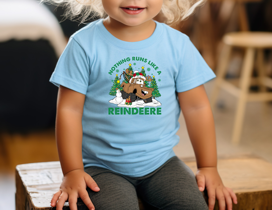 Toddler in light-blue Reindeere Baby Tee with tractor-reindeer graphic, sitting on a wooden bench. The shirt is soft, shrink-resistant, and perfect for festive occasions.