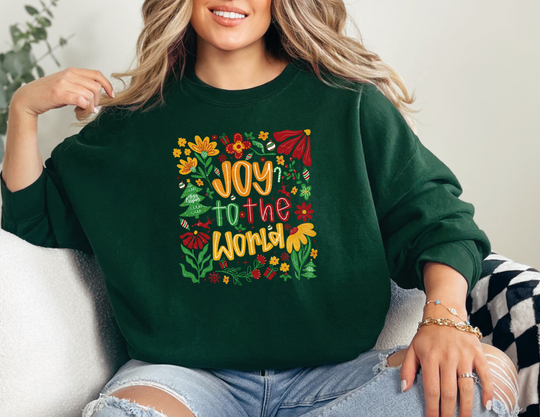 Woman in dark-green Joy to the World sweatshirt with festive motifs, wearing light-wash jeans and bracelets, showcasing cozy design by Faith Family Designs.