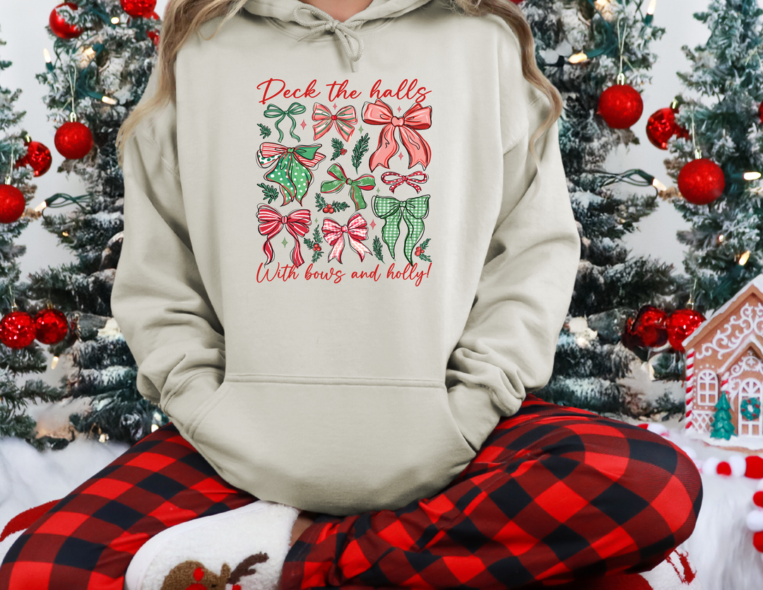 Person in buffalo-check pajama pants and white slippers wears a Deck The Halls beige hoodie with Christmas bows, seated in a festive setting.