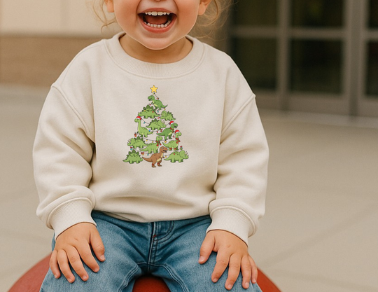 Toddler in light-cream Dino Tree Toddler Crew sweatshirt, featuring playful dinosaurs arranged as a Christmas tree, smiles while seated. Cozy, durable, and perfect for festive warmth.