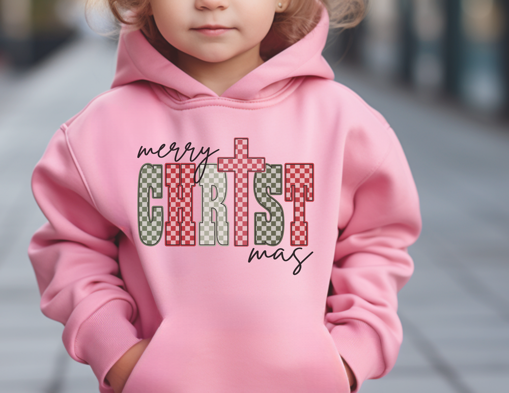 Child wearing Christmas Youth Hoodie with merry Christmas and checkerboard pattern lettering, standing outdoors. Hoodie features kangaroo pocket and ultra-soft fleece fabric.