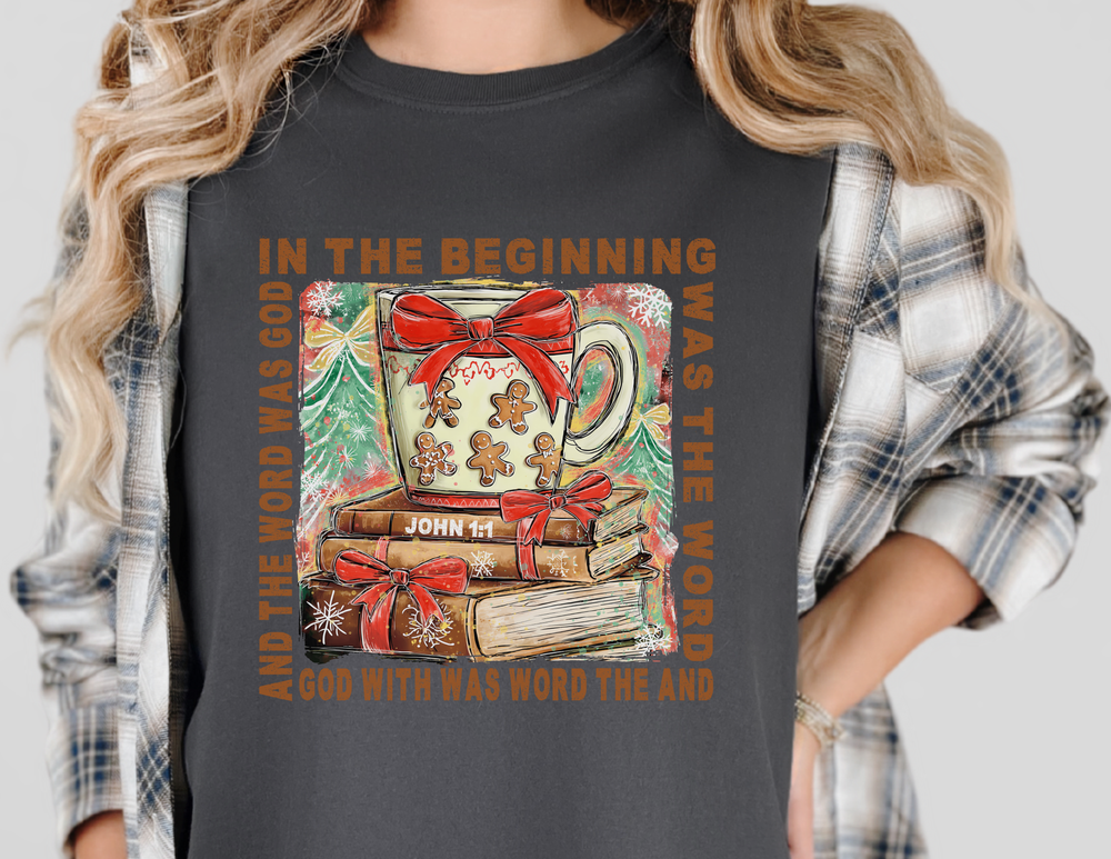 Person wearing In The Beginning Tee with festive Christmas illustration featuring a gingerbread-themed mug, ribbon-bound books, snowflakes, tree, and John 1:1 Bible verse.