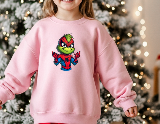 Young child wearing oversized Christmas Spider Toddler Crew sweatshirt, featuring Grinch as Spider-Man, stands by a decorated Christmas tree, evoking a festive and playful atmosphere.