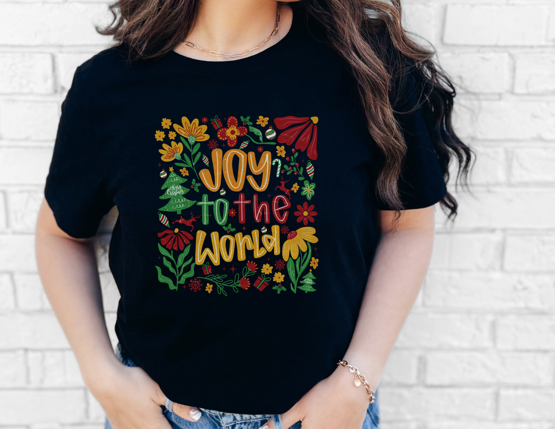 Person wearing Joy to the World Tee with festive motifs, standing against a white brick wall. The shirt is a soft, unisex, black jersey short sleeve.