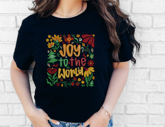 Person wearing Joy to the World Tee with festive motifs, standing against a white brick wall. The shirt is a soft, unisex, black jersey short sleeve.