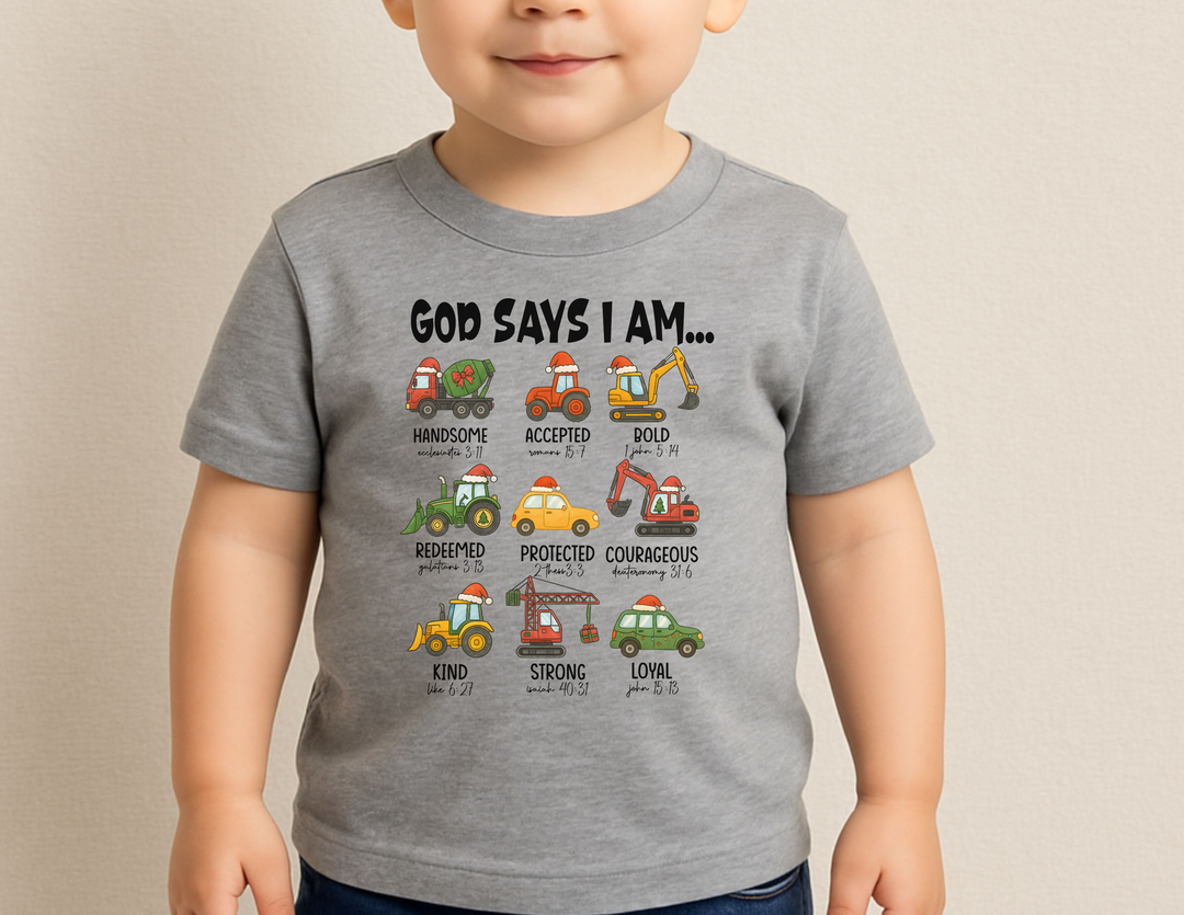 Young child wearing God Says Christmas Truck Baby Tee featuring cartoon construction vehicles with Santa hats and positive words, designed for comfort and durability.