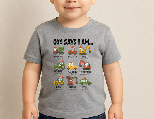 Young child wearing God Says Christmas Truck Baby Tee featuring cartoon construction vehicles with Santa hats and positive words, designed for comfort and durability.