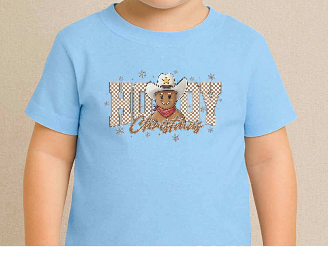 Child wearing Howdy Christmas Kids Tee with a cowboy gingerbread design, snowflakes, and festive text. Ideal for everyday wear with durable, soft cotton fabric.