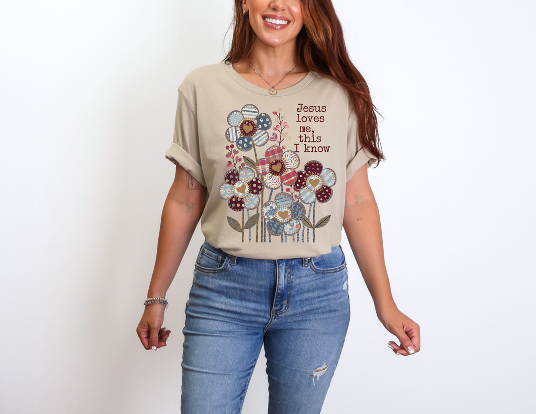 Person wearing a light-beige Jesus Loves Me Tee with patchwork floral design and phrase, paired with light-blue jeans, against a white background.