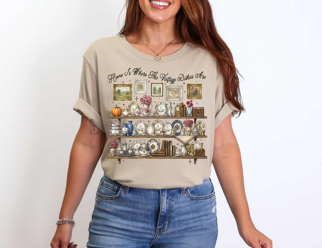 Vintage Dishes Tee featuring illustrated shelves of vintage china and decor, worn by a smiling person in cuffed sleeves and jeans, highlighting comfort and style.