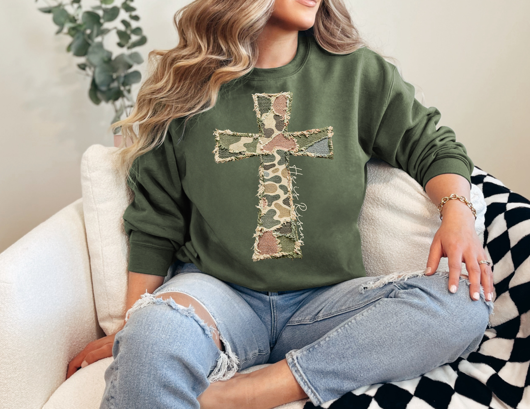 Person relaxing in a light armchair, wearing the He is Risen Cross Crew sweatshirt with a frayed camouflage cross design.