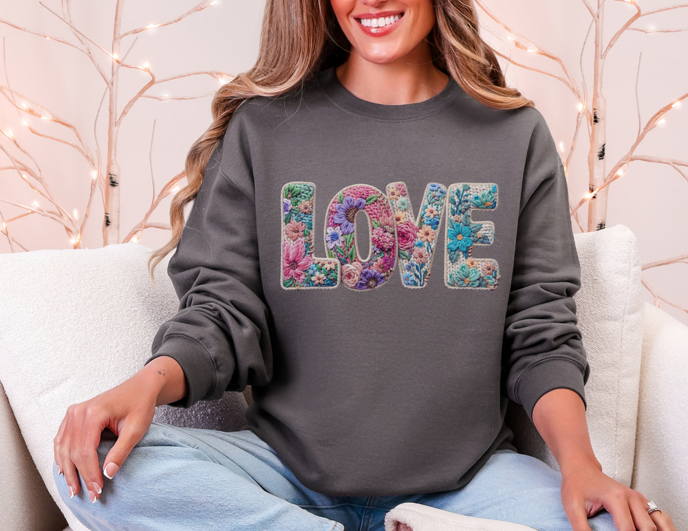 Person on a light couch wearing a dark gray Love Flower Crew sweatshirt, featuring floral embroidery on LOVE letters. Hands rest on lap, fairy-lit branches behind.