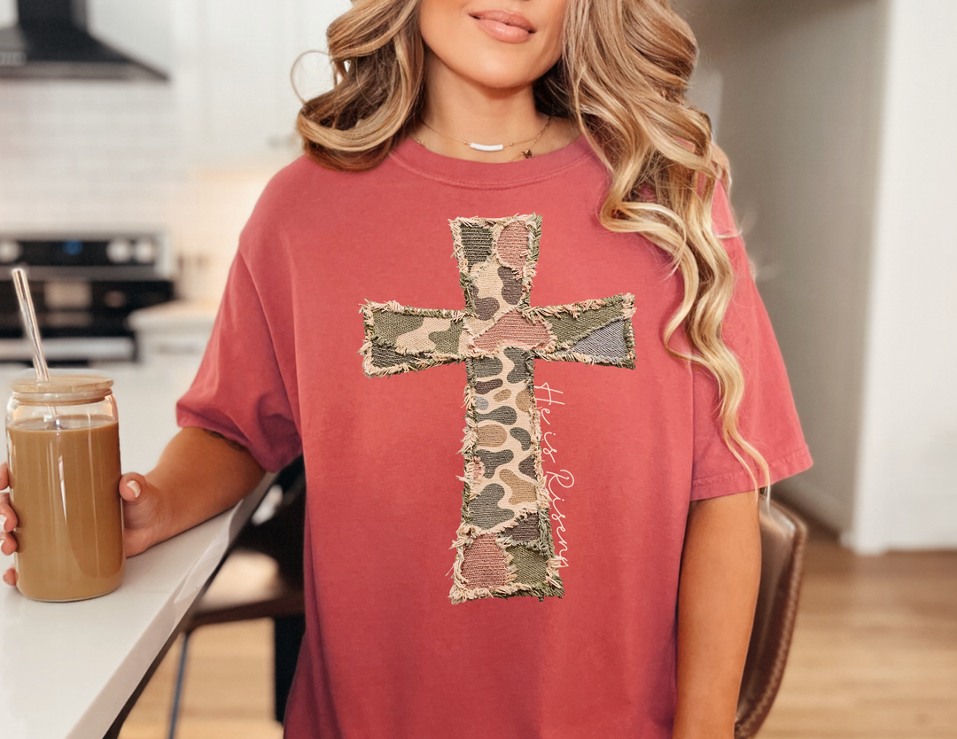 Person wearing He is Risen Distressed Tee with patchwork cross, holding a mason jar with iced coffee, standing in a bright kitchen.