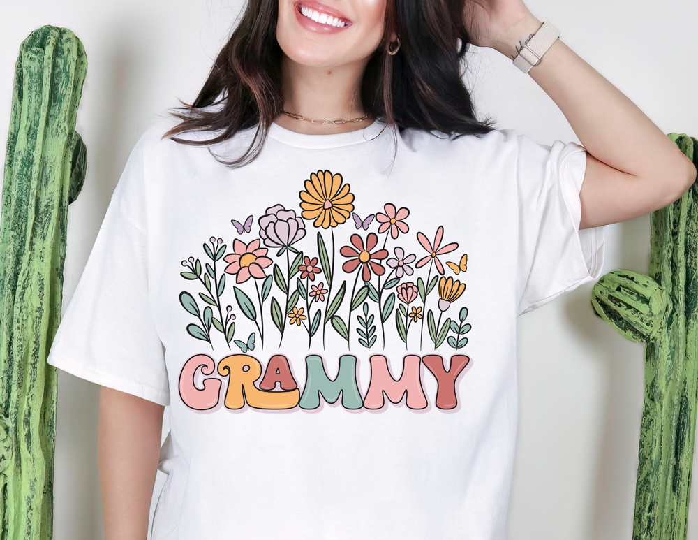 Person wearing Grammy Flower Tee with pastel flowers and butterflies, smiling in front of cactus plants. Classic unisex fit, soft cotton, perfect for everyday wear.