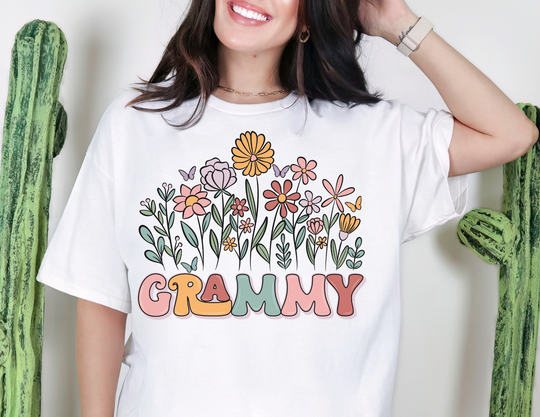 Person wearing Grammy Flower Tee with pastel flowers and butterflies, smiling in front of cactus plants. Classic unisex fit, soft cotton, perfect for everyday wear.