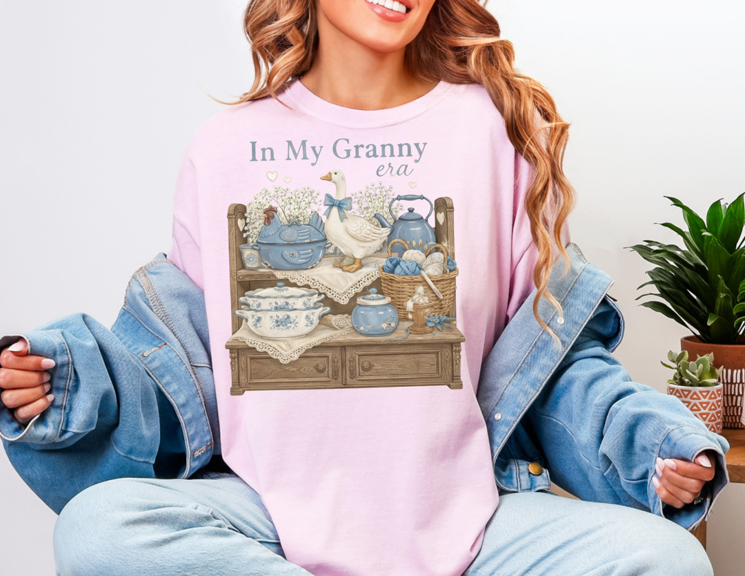 Person wearing In My Granny Era Tee with vintage motif on pastel pink sweatshirt, featuring cozy shelf items like a teapot, yarn, and flowers.