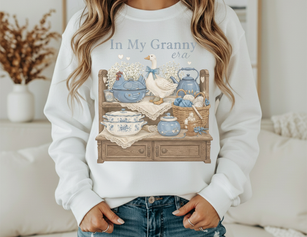 In My Granny Era Crew sweatshirt with vintage motif features a ceramic hen, baby’s-breath, and yarn on a hutch, embodying cozy nostalgia.