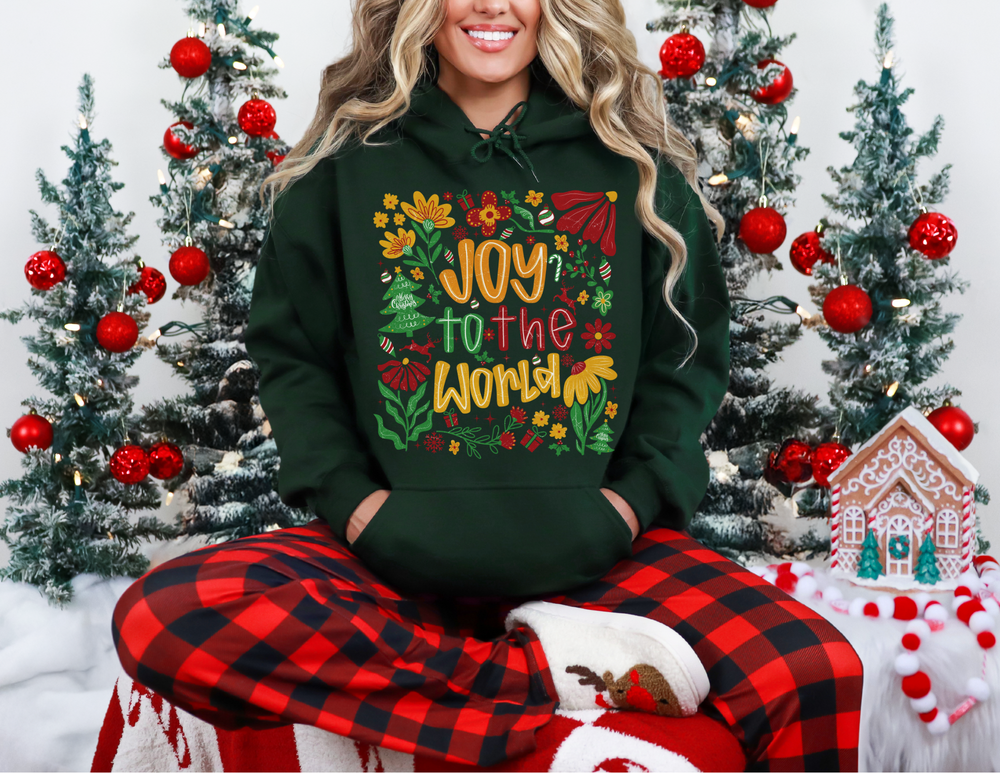 Person in buffalo-plaid pants and reindeer slippers wears the Joy to the World hoodie in a festive setting with decorated trees and a gingerbread house.