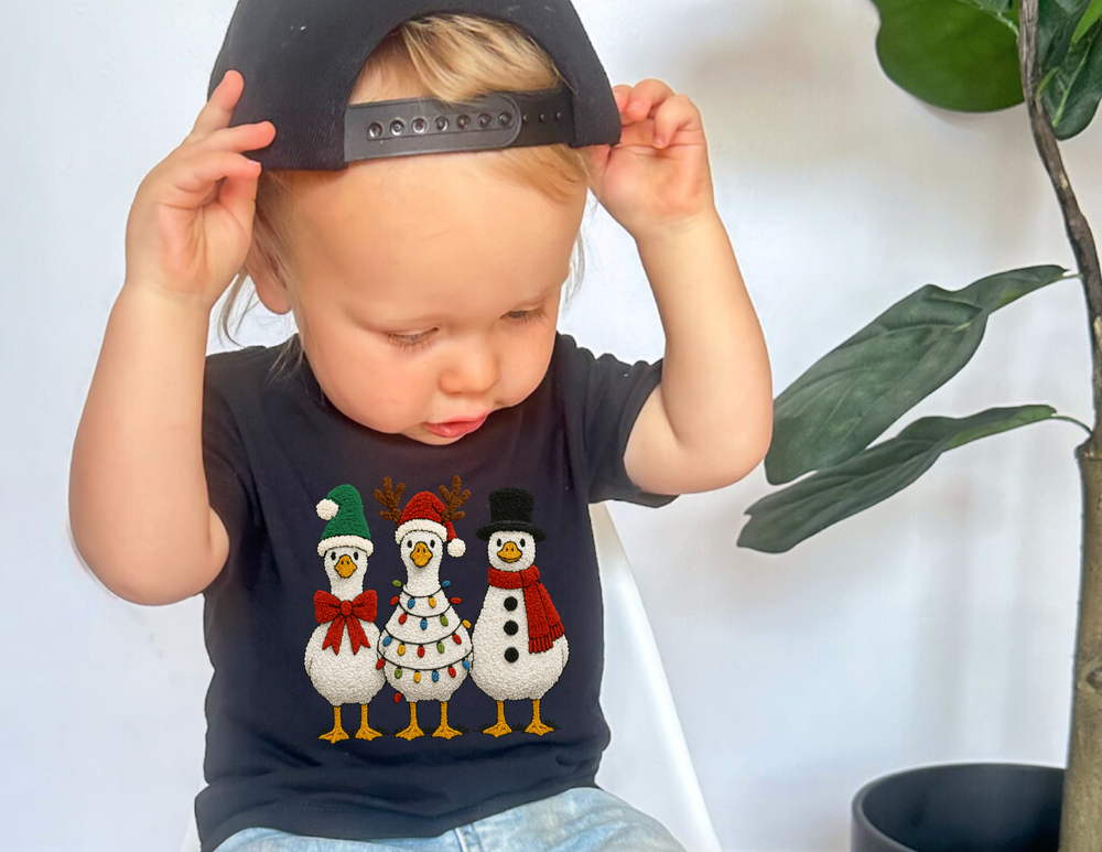 Toddler in a Christmas Geese Baby Tee, adjusting a baseball cap, featuring festive birds, exemplifying Faith Family Designs' playful and durable children's apparel.