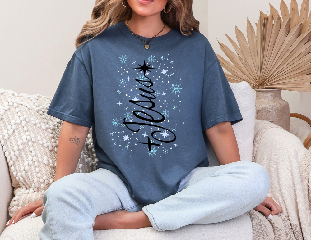 Woman in “Joyful” Jesus Tree Tee, blue with snowflakes, sitting on sofa, wearing light jeans; embodies cozy, casual winter style from Faith Family Designs.