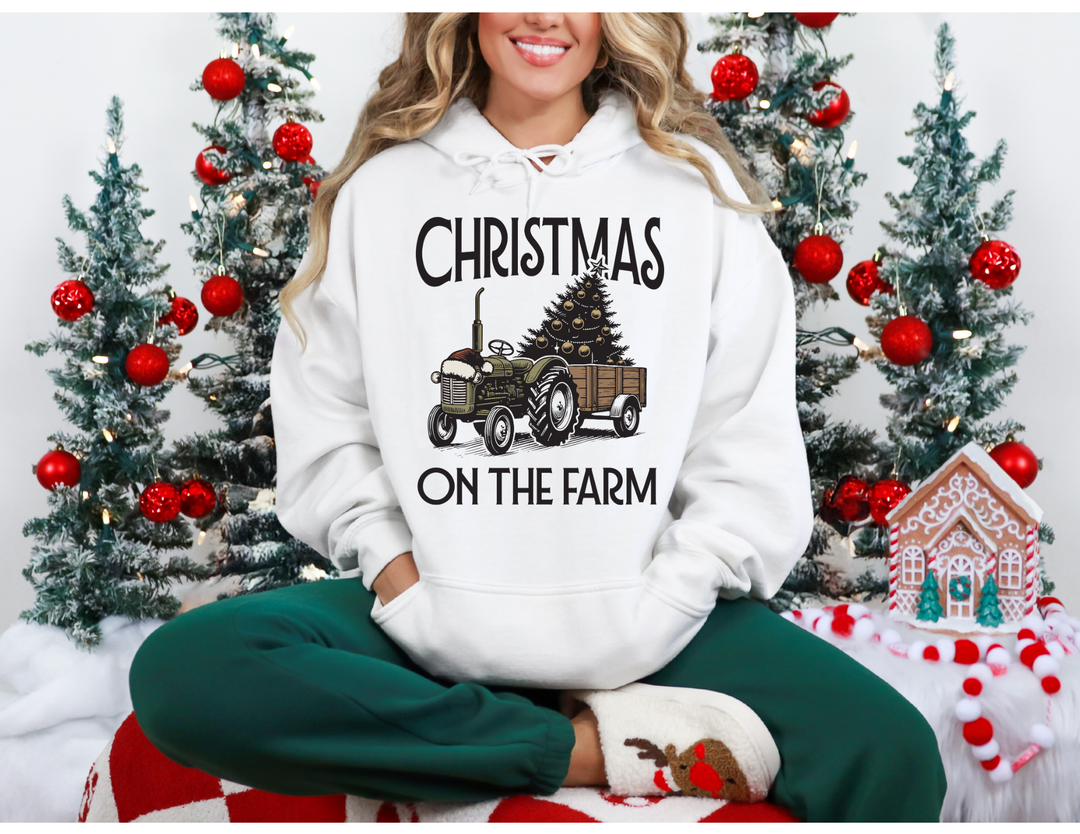 Person in Christmas on the Farm hoodie, featuring a tractor and tree, in a cozy holiday setting with Christmas trees and gingerbread decor.
