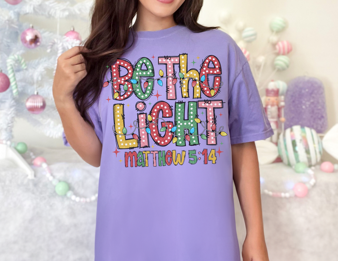 Person wearing a Be the Light oversized lavender T-shirt featuring multicolored lettering and holiday-themed designs.