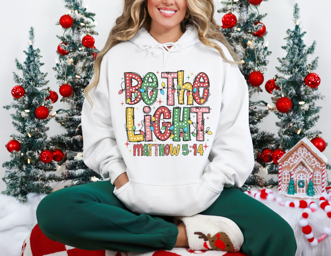 Person in a Be the Light hoodie, seated amid festive decor, showcasing a cozy, holiday-themed sweatshirt perfect for warmth and style.