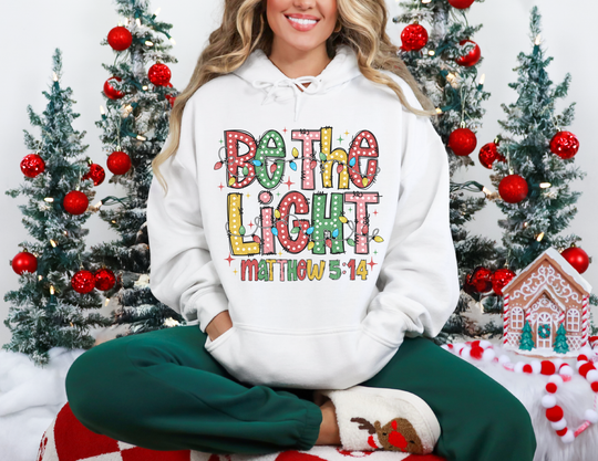 Person in a Be the Light hoodie, seated amid festive decor, showcasing a cozy, holiday-themed sweatshirt perfect for warmth and style.