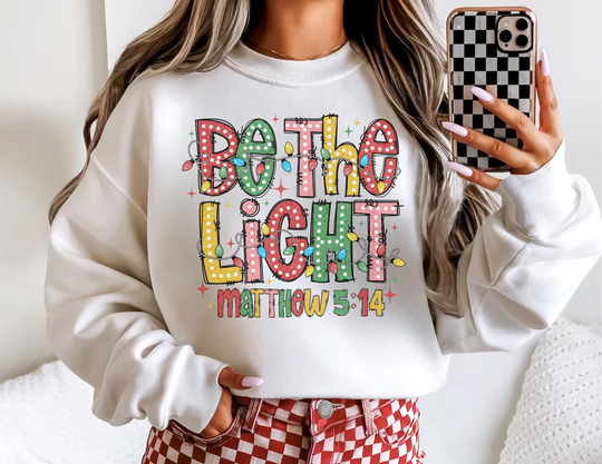 Person wearing Be the Light Crew sweatshirt with festive lettering and Christmas lights, paired with checkerboard phone case and pants, showcasing casual, cozy style.
