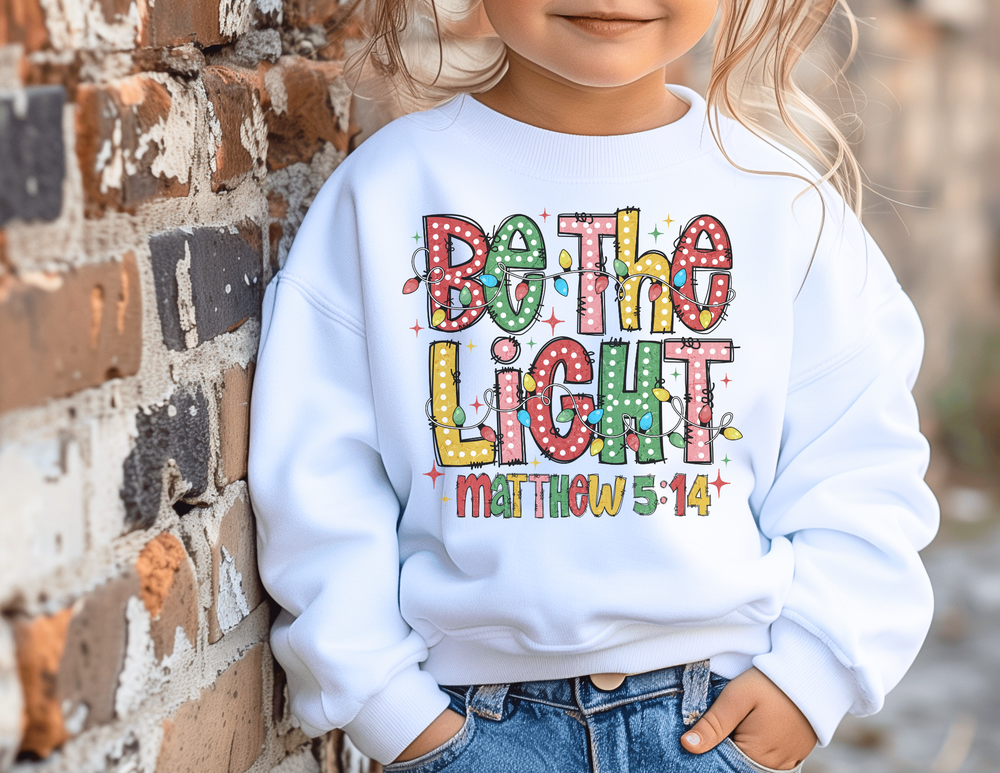 Child wearing Be the Light Toddler Crew sweatshirt with festive design, hands in jeans pockets, against a brick wall. Cozy fleece blend for warmth and comfort.