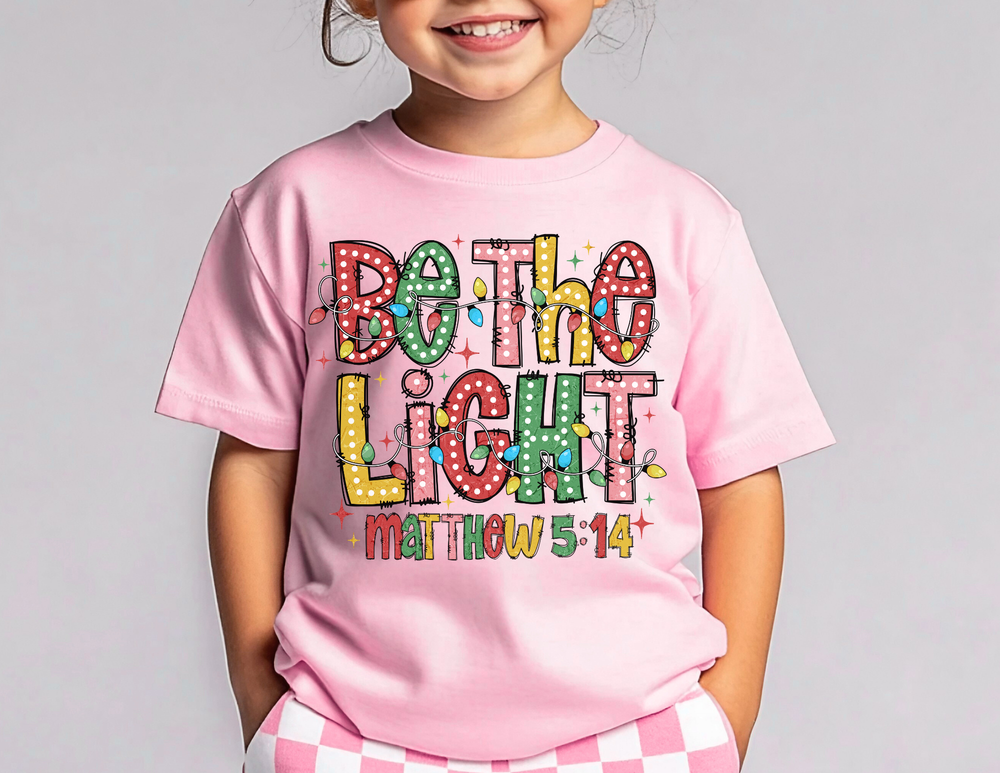 Child wearing a pale-pink Be the Light kids tee with Christmas-style lights and scripture reference, paired with pink-and-white checkered bottoms, smiling.