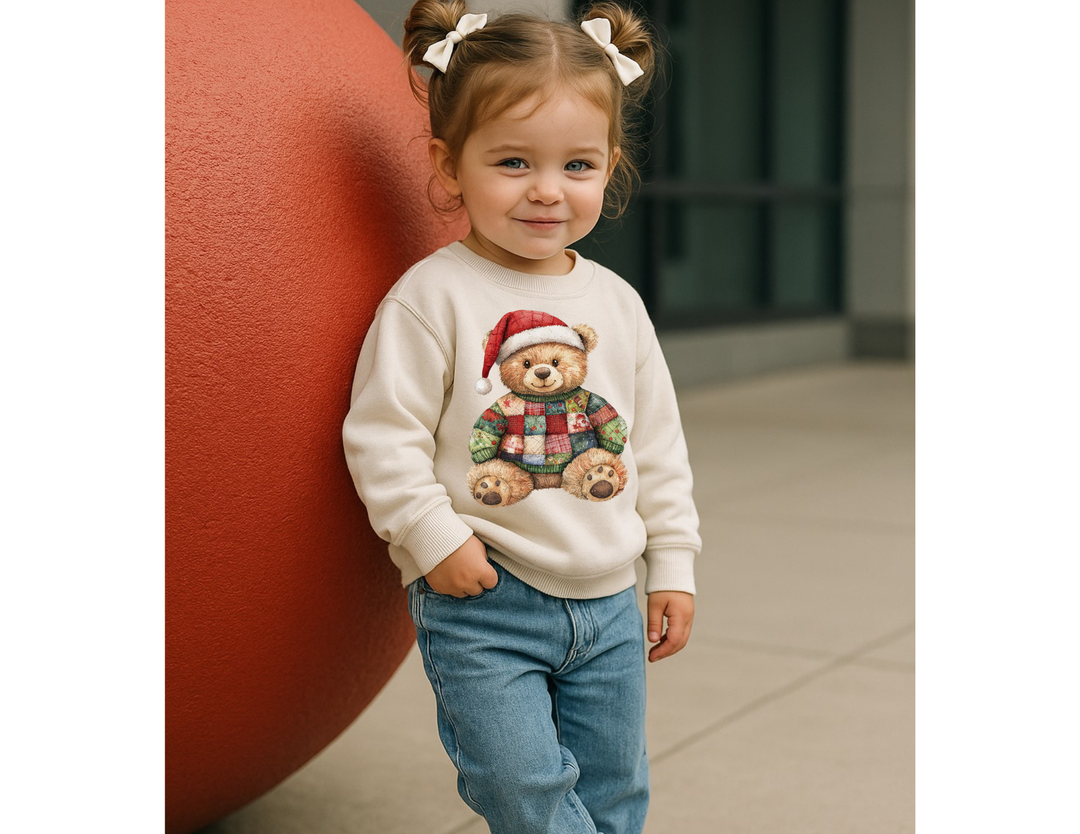 Toddler in Christmas Bear Toddler Crew sweatshirt, featuring a patchwork teddy bear in a Santa hat, stands against a red sphere, smiling sweetly with hair in buns.